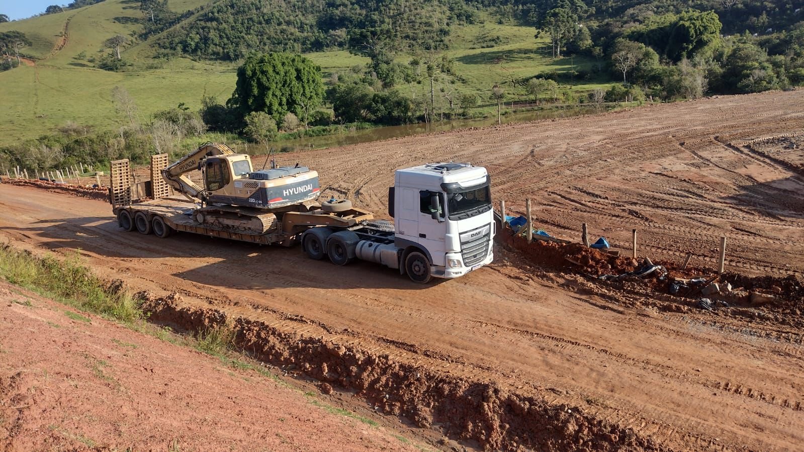 Caminhão DAF com carreta prancha rebaixada transportando escavadeira Hyundai em obra de terraplenagem